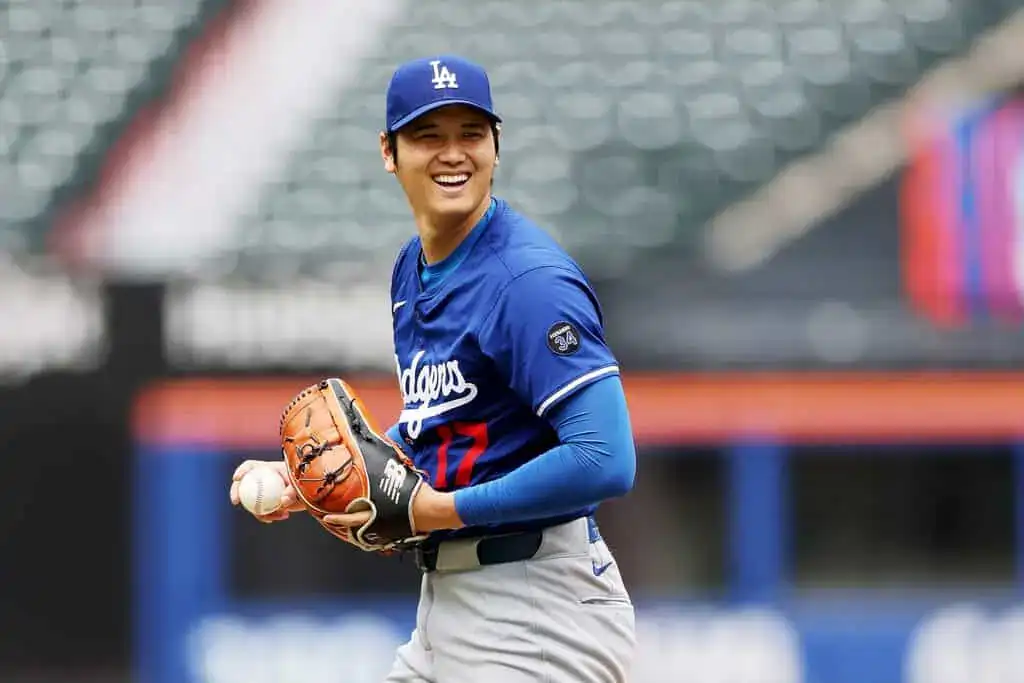 A pitcher in a blue Dodgers jersey grips a baseball, preparing to throw, with a baseball field setting in the background.