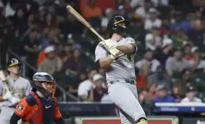 A baseball player in a grey uniform swings a bat, as a catcher in orange gear prepares for the play, amid a cheering crowd.