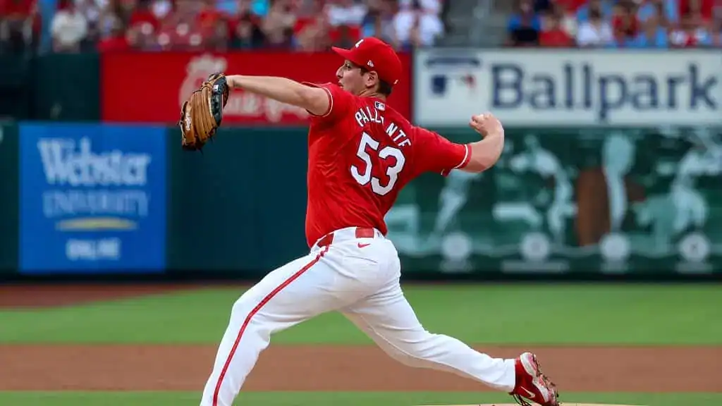 A baseball pitcher in a bright red uniform winds up to throw the ball on a lively field, with fans in the stands behind him.