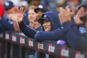 A man in a blue hoodie is leaning on a fence. He is wearing a baseball cap and appears to be outdoors, possibly at an event with a crowd around him.