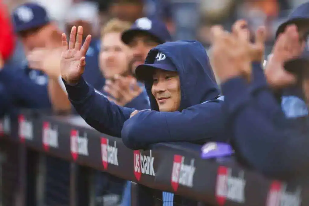 A man in a blue hoodie is leaning on a fence. He is wearing a baseball cap and appears to be outdoors, possibly at an event with a crowd around him.