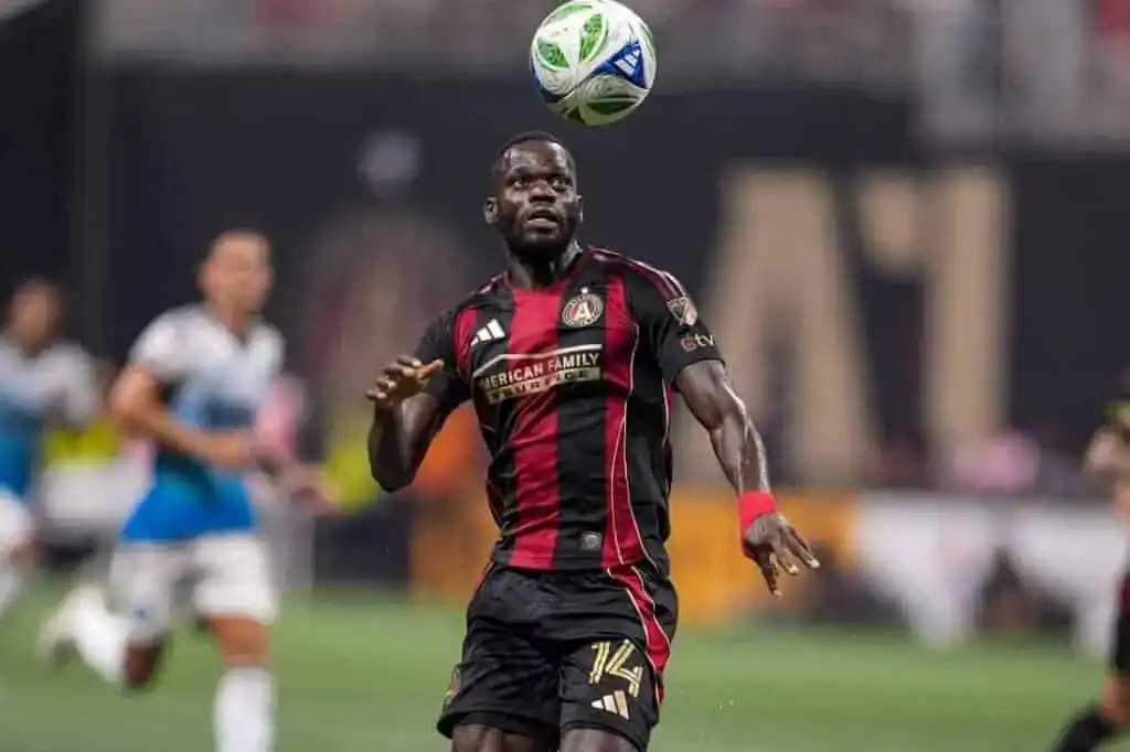 A man is depicted in a football uniform, holding a ball in his hand. The setting appears to be outdoors on grass, likely during an athletic game.