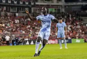 A soccer player in a light blue jersey celebrates on the field, with fans cheering in the background during a lively match.