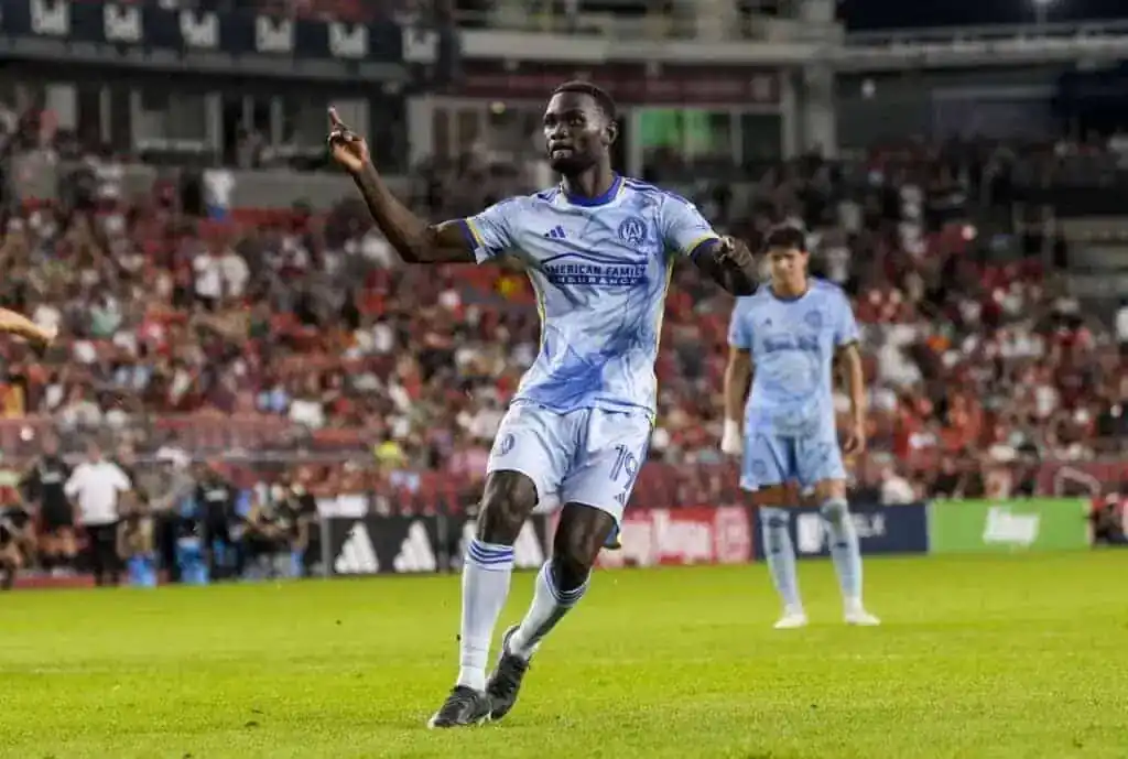 A soccer player in a light blue jersey celebrates on the field, with fans cheering in the background during a lively match.
