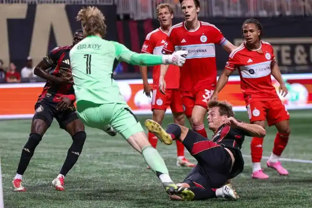 A goalkeeper dives to stop the ball as a player falls during an intense soccer match, surrounded by teammates and opponents.