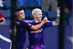 Two soccer players in purple jerseys celebrate a victory, one raising their fist in triumph while the other embraces them.