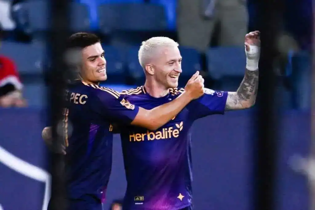 Two soccer players in purple jerseys celebrate a victory, one raising their fist in triumph while the other embraces them.