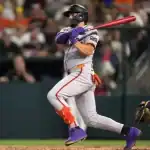 A baseball player in a gray and orange uniform swings a bat during a game, with a catcher in green gear ready to receive the pitch.