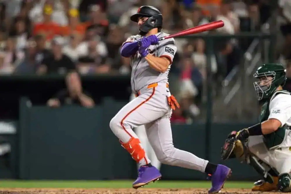 A baseball player in a gray and orange uniform swings a bat during a game, with a catcher in green gear ready to receive the pitch.