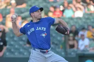 A male pitcher in a blue Blue Jays jersey winds up to throw a baseball, with a blurred crowd in the background.