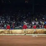 Grounds crew in red shirts rolls out a tarp on the baseball field as a crowd watches from the stadium seats in the background.
