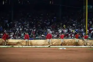 Grounds crew in red shirts rolls out a tarp on the baseball field as a crowd watches from the stadium seats in the background.