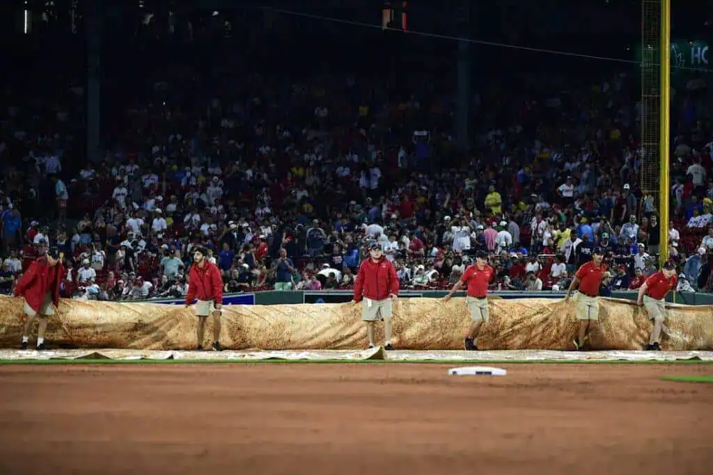 Grounds crew in red shirts rolls out a tarp on the baseball field as a crowd watches from the stadium seats in the background.