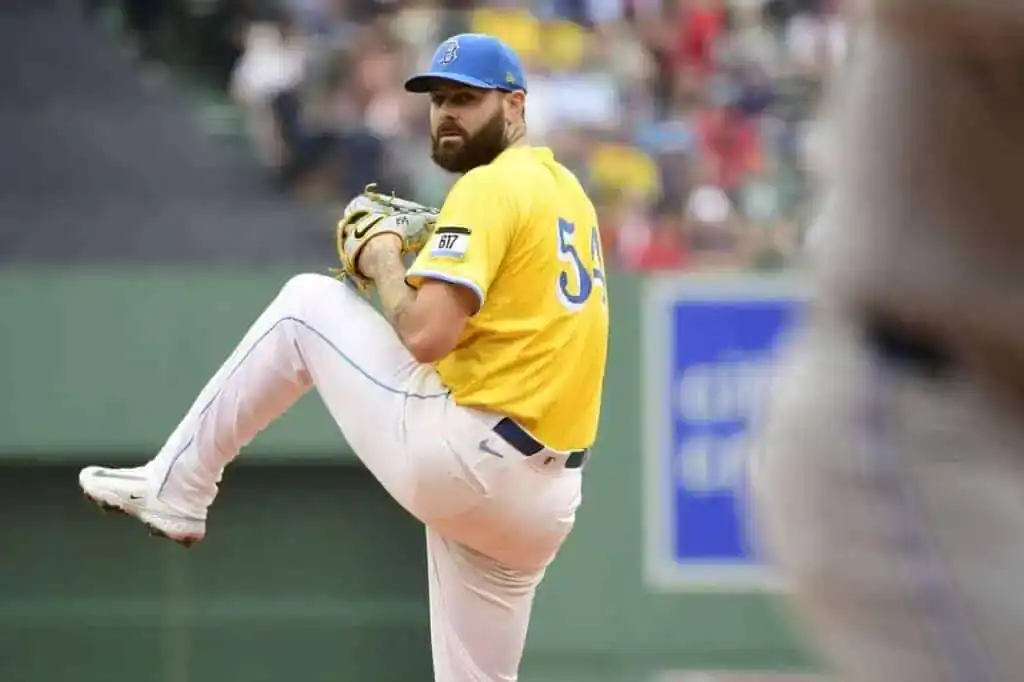 A baseball pitcher in a yellow jersey and blue cap winds up to throw, showcasing a tense moment during the game with a blurred crowd in the background.