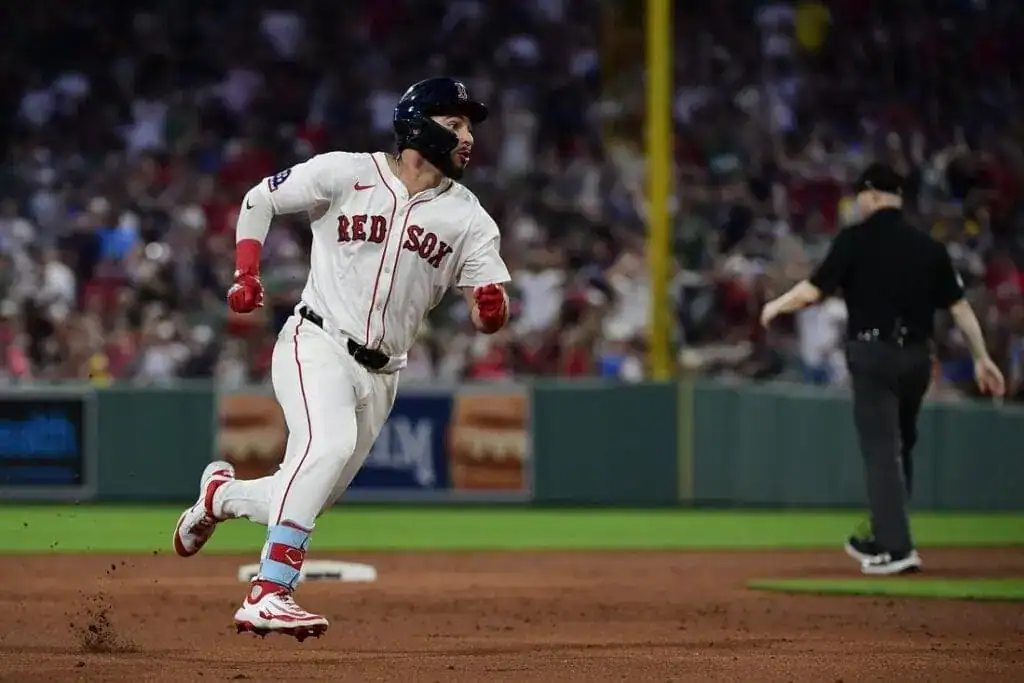 A Boston Red Sox player sprints towards base as the crowd cheers in the background, with an umpire observing the play.