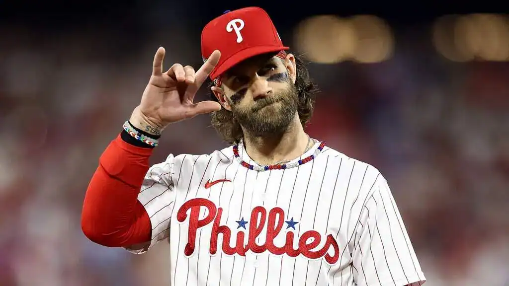 Philadelphia Phillies player Bryce Harper in a pinstripe jersey and red cap makes a hand gesture.