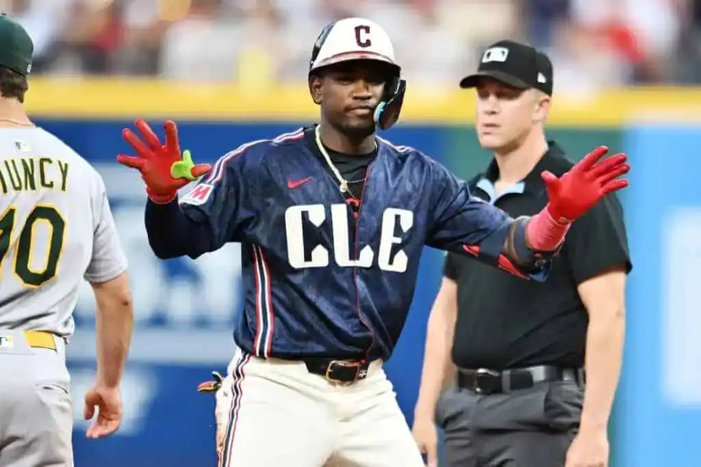 A Cleveland Guardians player in a blue "CLE" City Connect jersey stands on base and gestures with his hands.