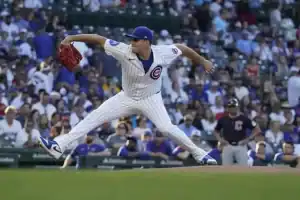 A baseball pitcher in a Cubs uniform delivers a pitch, with a lively crowd in blue and white jerseys cheering in the background.