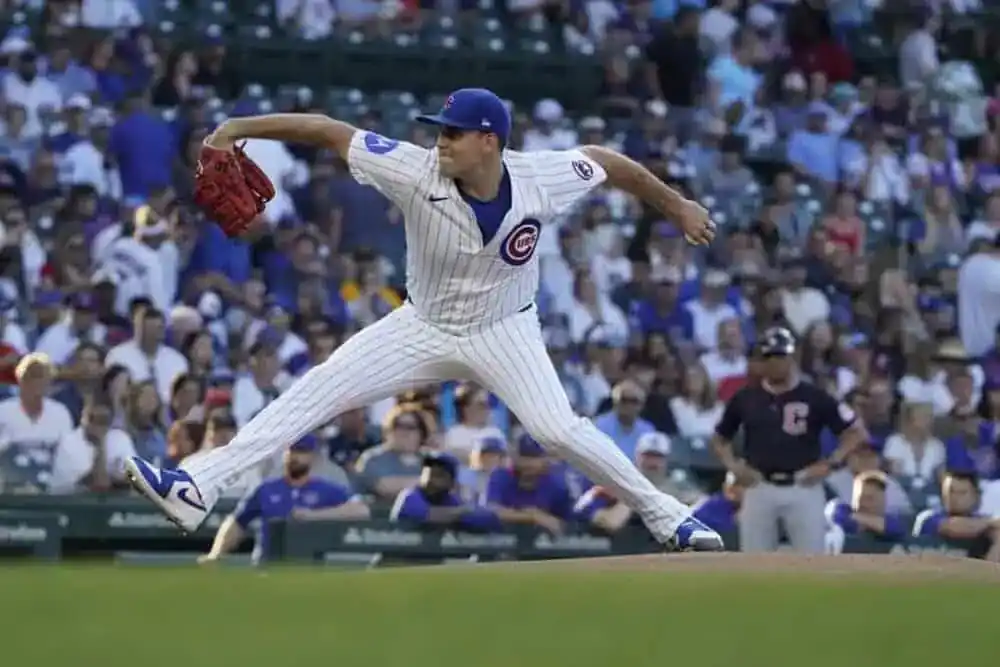 A baseball pitcher in a Cubs uniform delivers a pitch, with a lively crowd in blue and white jerseys cheering in the background.