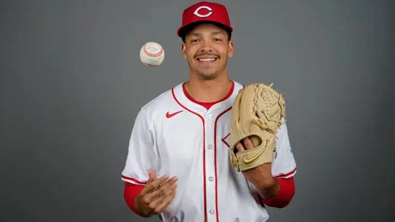 A baseball player wearing a white jersey with red accents and a red cap holds a baseball and glove, poised as if to throw.