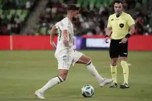 A soccer player in a white uniform dribbles the ball on the field while a referee in a yellow shirt observes the play.