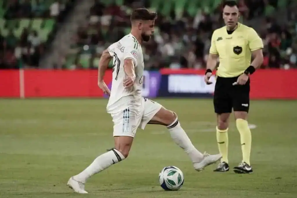 A soccer player in a white uniform dribbles the ball on the field while a referee in a yellow shirt observes the play.