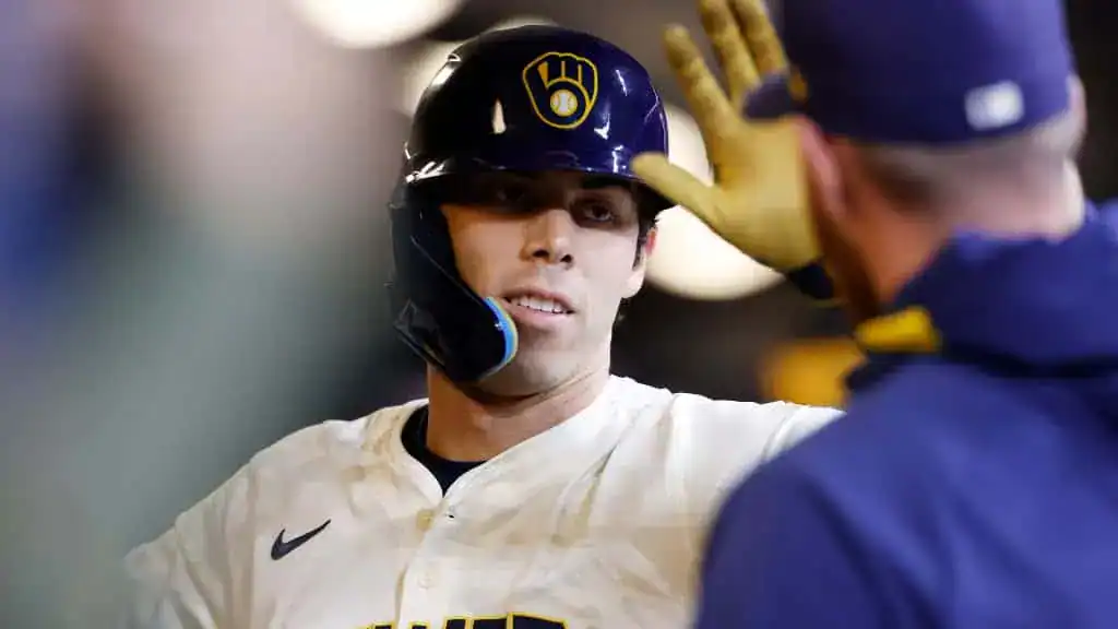 A baseball player in a white jersey celebrates with a coach in a blue cap, exchanging high-fives in a lively dugout atmosphere.