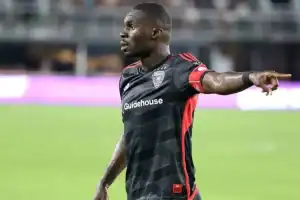 A man in a football uniform is depicted in the image, pointing his arm. The setting appears to be an outdoor soccer field, likely within a soccer-specific stadium