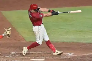 A baseball player in a red uniform swings his bat at a pitch, while a catcher crouches behind him in anticipation of the play.