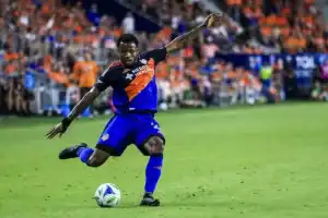 A soccer player in a blue and orange uniform skillfully strikes a ball on a vibrant field, surrounded by cheering fans.