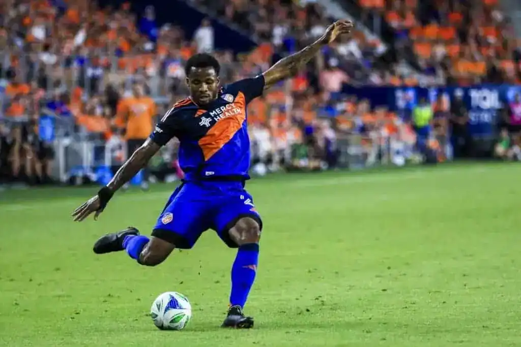 A soccer player in a blue and orange uniform skillfully strikes a ball on a vibrant field, surrounded by cheering fans.