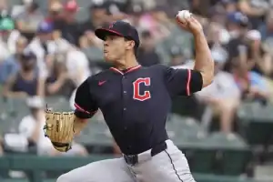 A baseball pitcher in a black jersey with a red 'C' logo prepares to throw a ball on a sunny day, surrounded by fans in the stands.