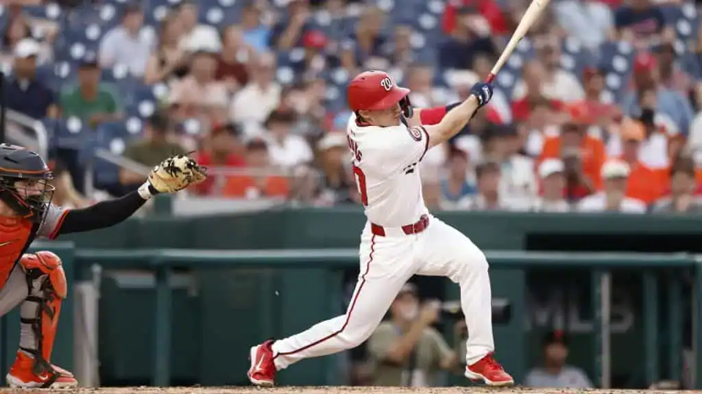 A batter, wearing a white and red uniform, is in mid-swing, with the bat connected to a ball.
