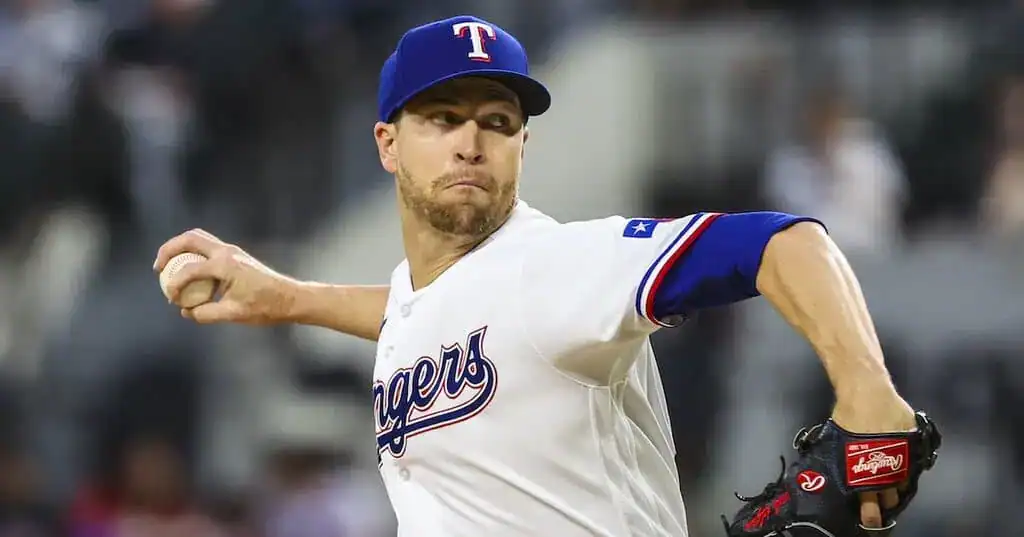 A Texas Rangers pitcher in action, throwing a baseball while wearing a team jersey and cap, against a blurred stadium background.