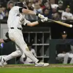 A baseball player swings a bat in a dimly lit stadium, with a catcher poised to receive the pitch behind him. Fans watch from the stands.