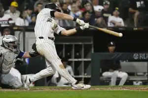 A baseball player swings a bat in a dimly lit stadium, with a catcher poised to receive the pitch behind him. Fans watch from the stands.