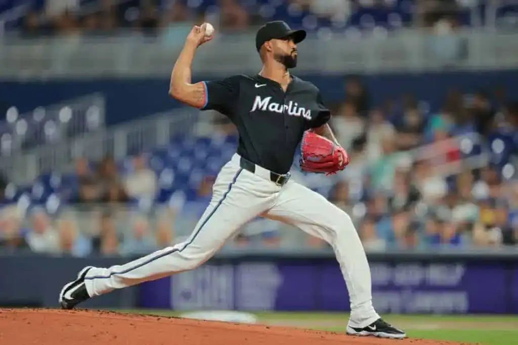 A pitcher in a black Marlins jersey prepares to throw a baseball on the mound, with fans visible in the blurred background.