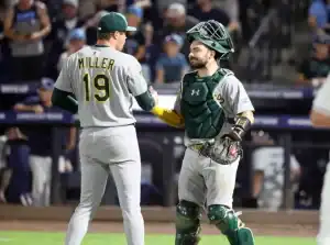 A player in a gray baseball uniform with "MILLER" and number 19 converses with a catcher in gear, set against a stadium backdrop.