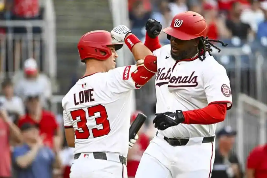 Two baseball players in red and white jerseys celebrate with fist bumps on the field, with fans blurred in the background.