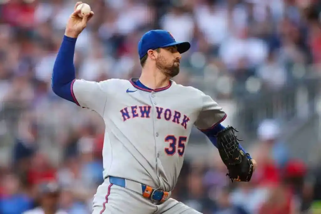 A New York Mets pitcher in a gray uniform delivers a baseball on the field, with a blurred crowd in the background.