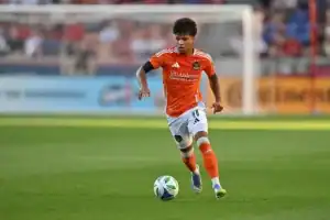 A soccer player in an orange jersey dribbles a ball across a green field during a sunny match, with a blurred crowd in the background.