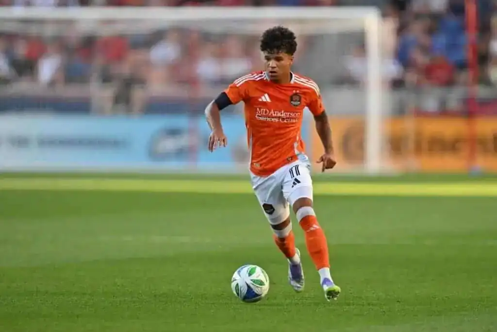 A soccer player in an orange jersey dribbles a ball across a green field during a sunny match, with a blurred crowd in the background.