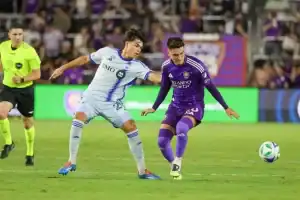 One wears a light blue uniform and the other a purple one. A referee stands in the background, observing the play.