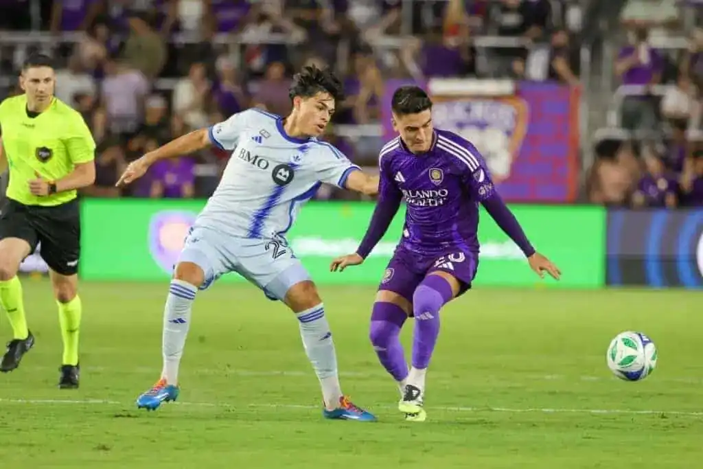 One wears a light blue uniform and the other a purple one. A referee stands in the background, observing the play.