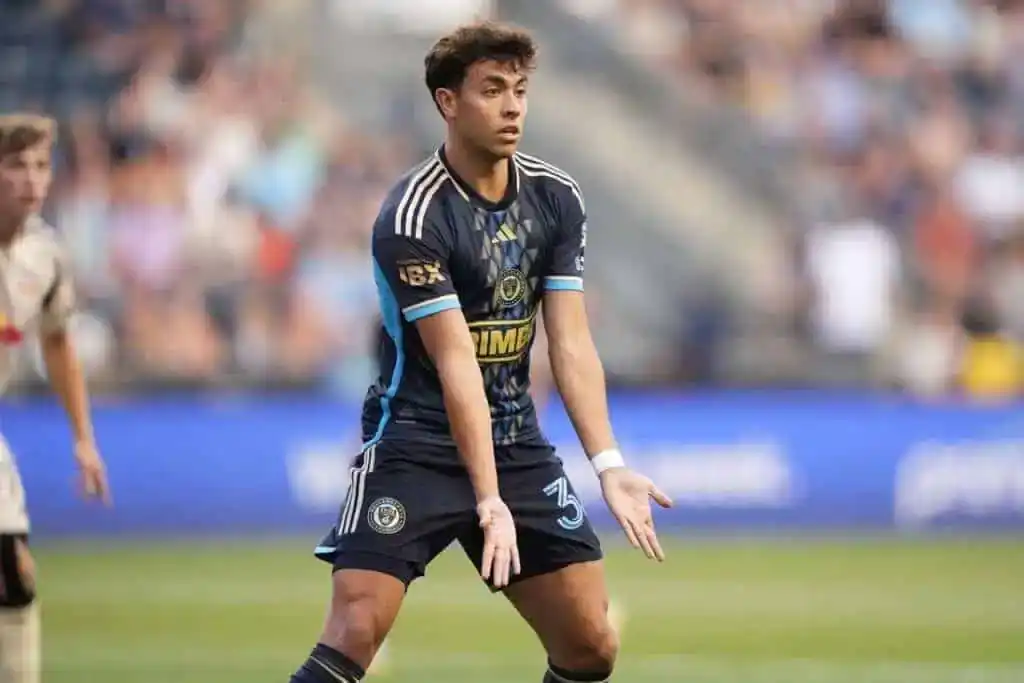 The image shows a soccer player, wearing a dark uniform with blue and gold details, standing on a soccer field.