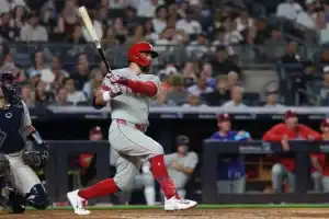 A player in a grey jersey swings a bat in a baseball game, with spectators and teammates visibly engaged in the background.