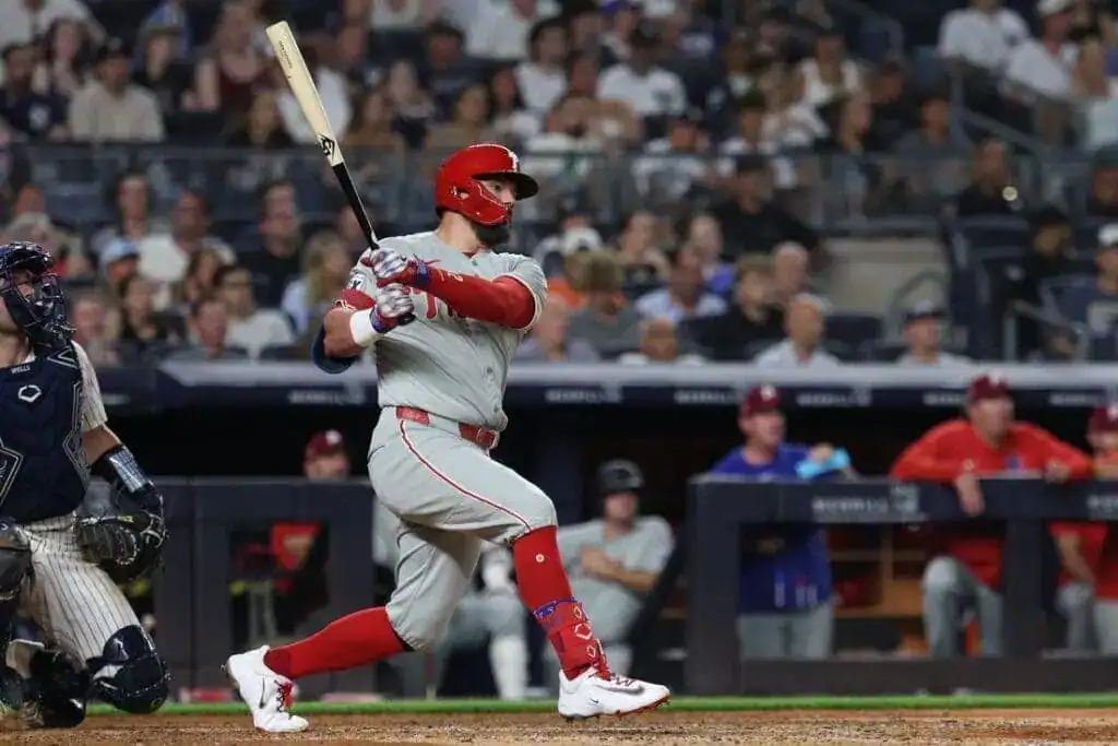 A player in a grey jersey swings a bat in a baseball game, with spectators and teammates visibly engaged in the background.