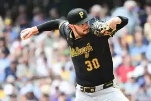 A Pittsburgh Pirates player winds up to pitch on the field, wearing a black jersey with yellow accents and surrounded by a lively crowd.