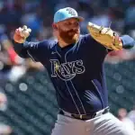 A baseball player in a navy uniform and light blue cap winds up to pitch on a sunny day at a crowded stadium.