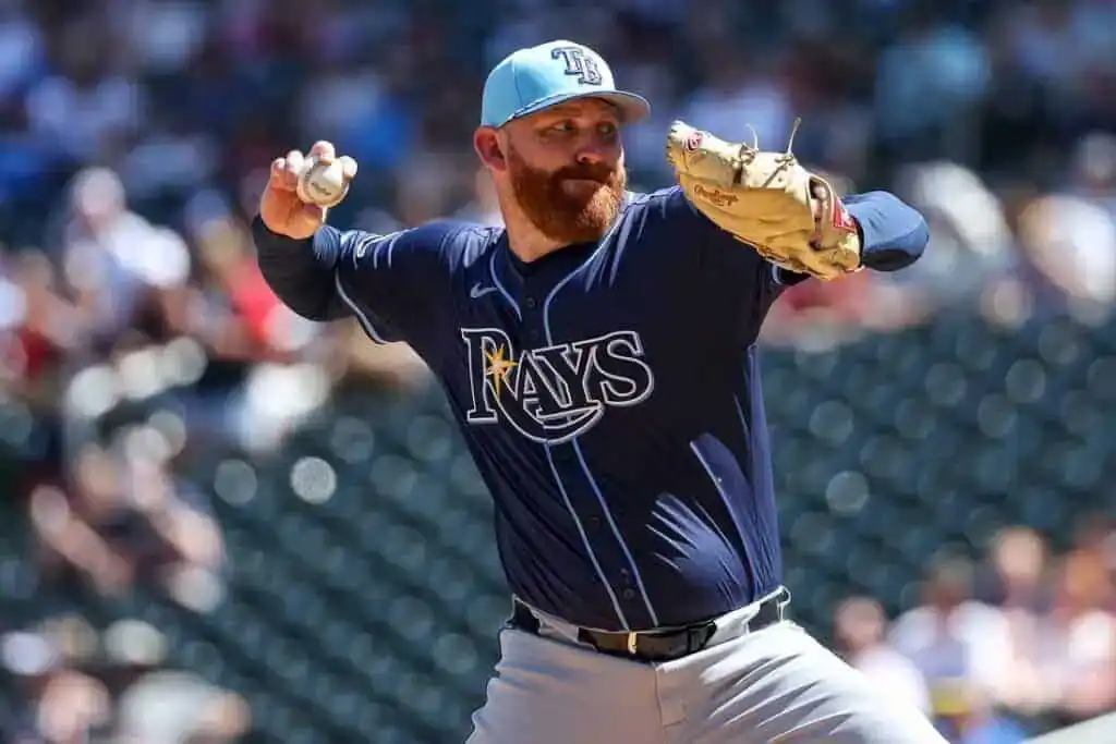 A baseball player in a navy uniform and light blue cap winds up to pitch on a sunny day at a crowded stadium.